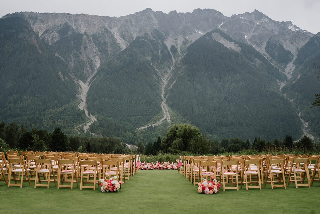 Rows of wooden chairs with pink floral arrangements set up outdoors on a lawn for a Big Sky Golf Wedding, facing a mountain range under a cloudy sky.
