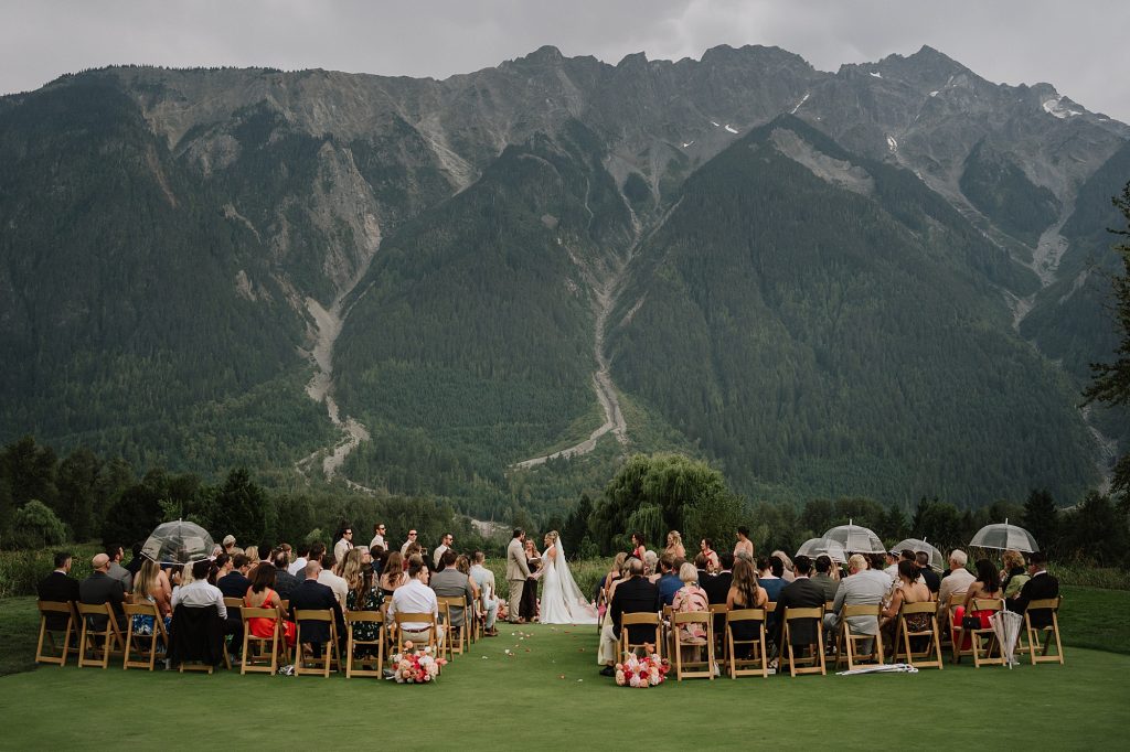 A Big Sky Golf Wedding ceremony unfolds outdoors with guests seated before a breathtaking mountain backdrop; the wedding party stands at the front as some guests hold transparent umbrellas.