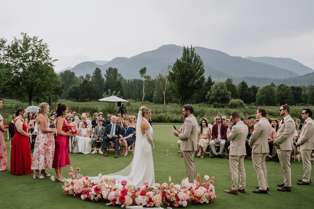 A bride and groom stand at an outdoor altar exchanging vows at their Big Sky Golf Wedding, surrounded by their wedding party, guests, and breathtaking mountains in the background.