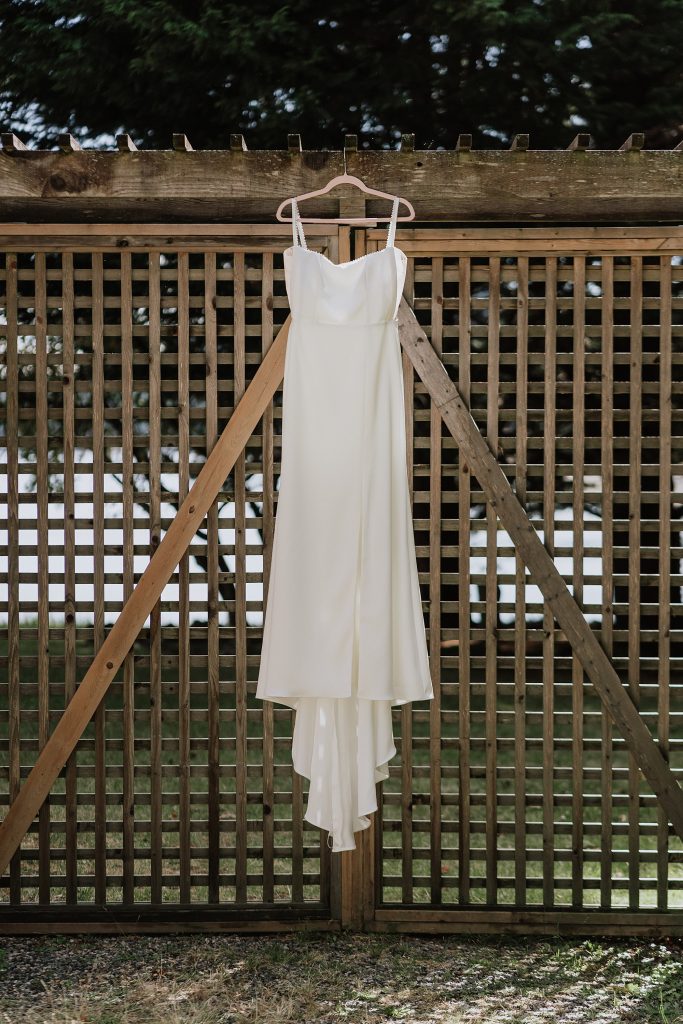 A simple white sleeveless dress hangs on a wooden lattice gate outdoors, with trees visible in the background.