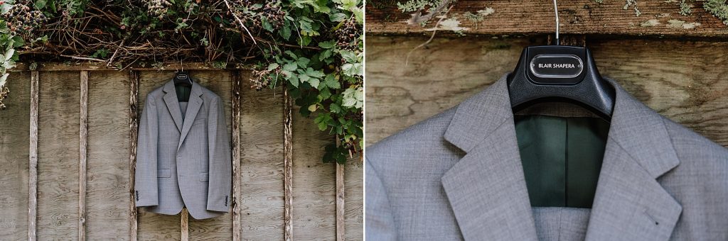 A light gray suit jacket hangs on a wooden wall under greenery and branches, displayed on a hanger labeled "Blair Shapera.