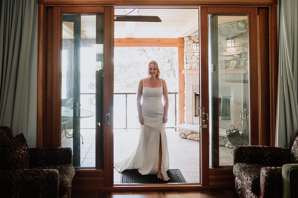 A woman in a white dress stands smiling in a doorway between a living area and a covered patio with stone walls and a fireplace.