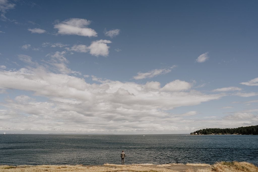 A person stands on a grassy shore facing a large body of water under a partly cloudy sky with distant land visible on the horizon.