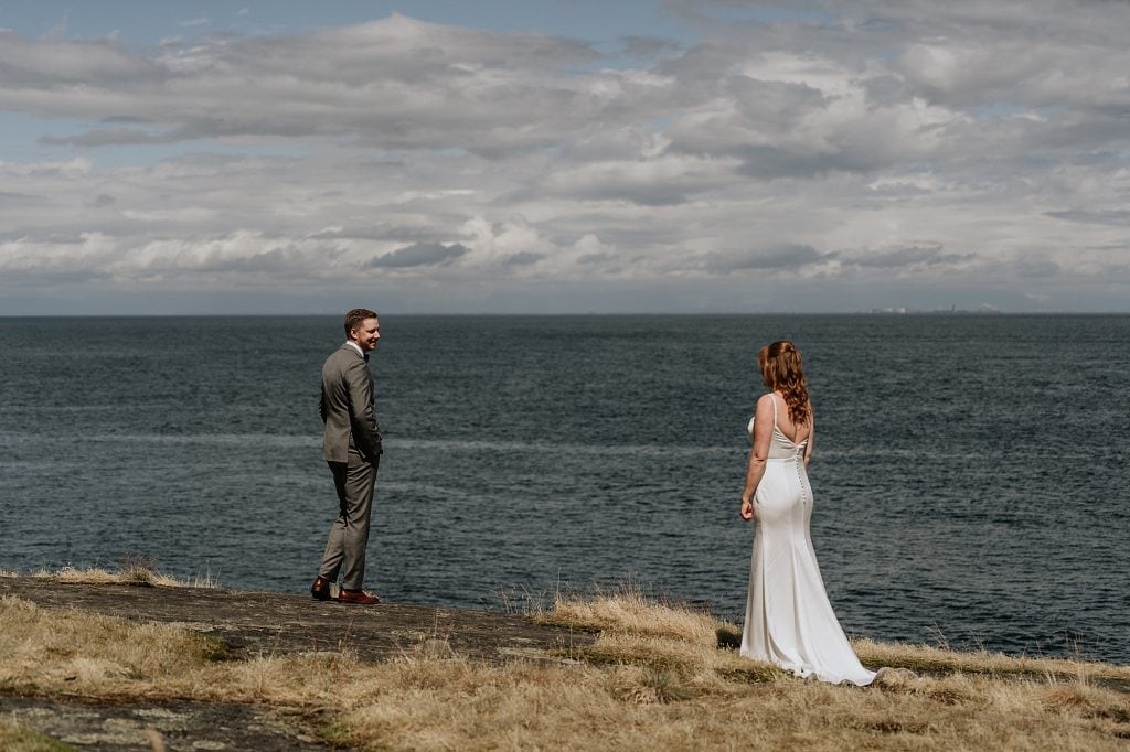 A man in a suit and a woman in a white dress stand facing each other on a grassy cliff overlooking the ocean under a cloudy sky.