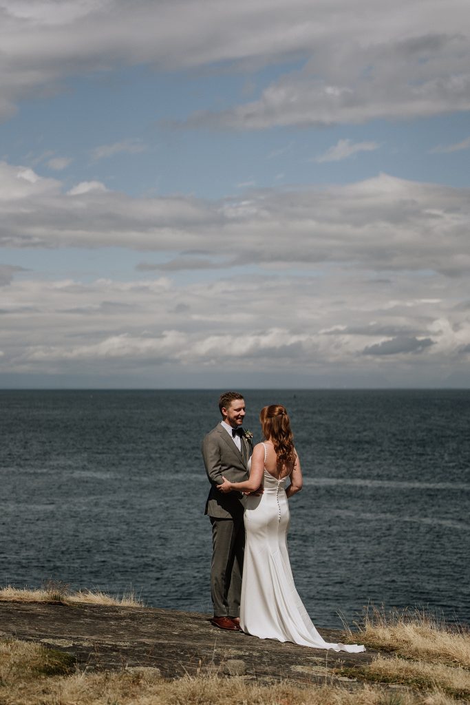 A couple dressed in wedding attire stands facing each other on a grassy cliff by the ocean, with cloudy skies above.