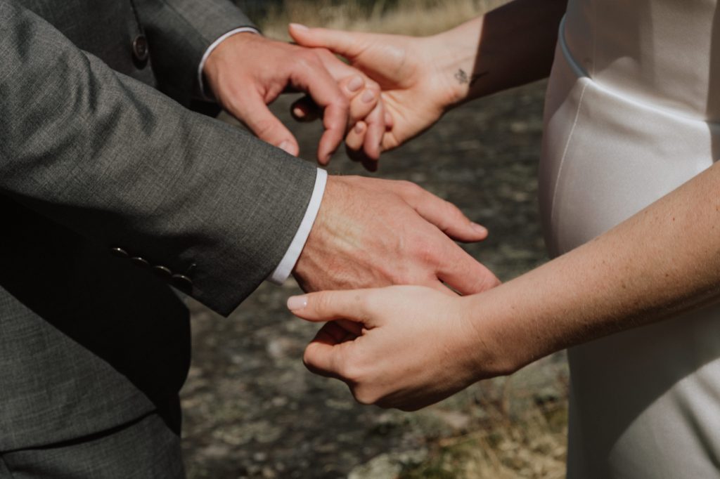 A close-up of two people holding hands, one wearing a gray suit and the other in a white dress, outdoors.