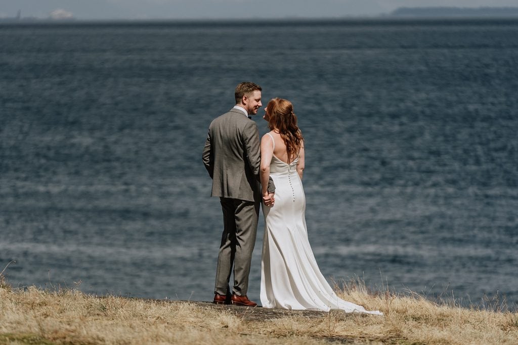 A bride and groom stand together on a grassy area facing a large body of water, both dressed in formal wedding attire.