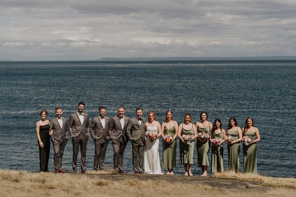 A wedding party poses outdoors on grass in front of a large body of water, with bridesmaids in green dresses and groomsmen in grey suits.