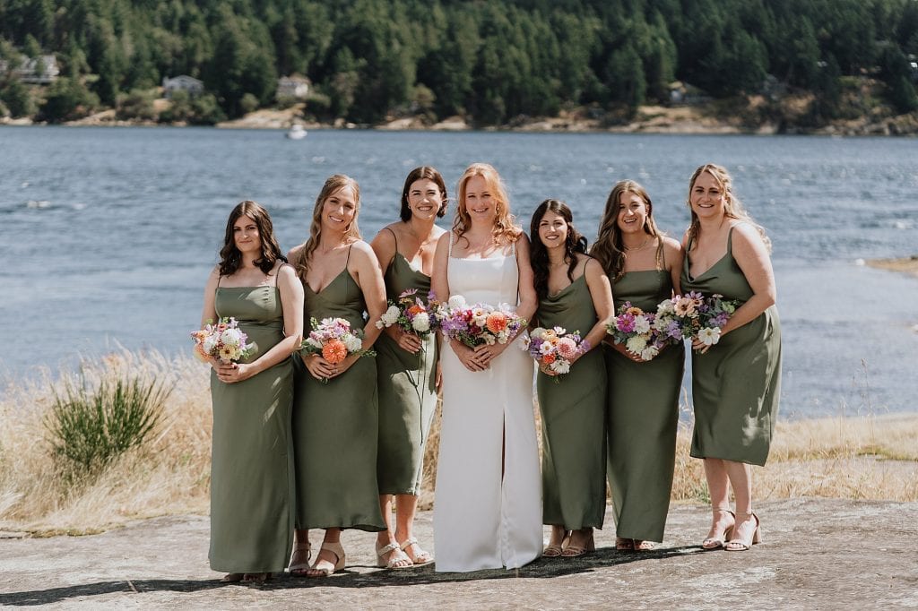A bride in a white dress stands with six bridesmaids in matching olive green dresses, all holding bouquets, with a lake and forested hills in the background.
