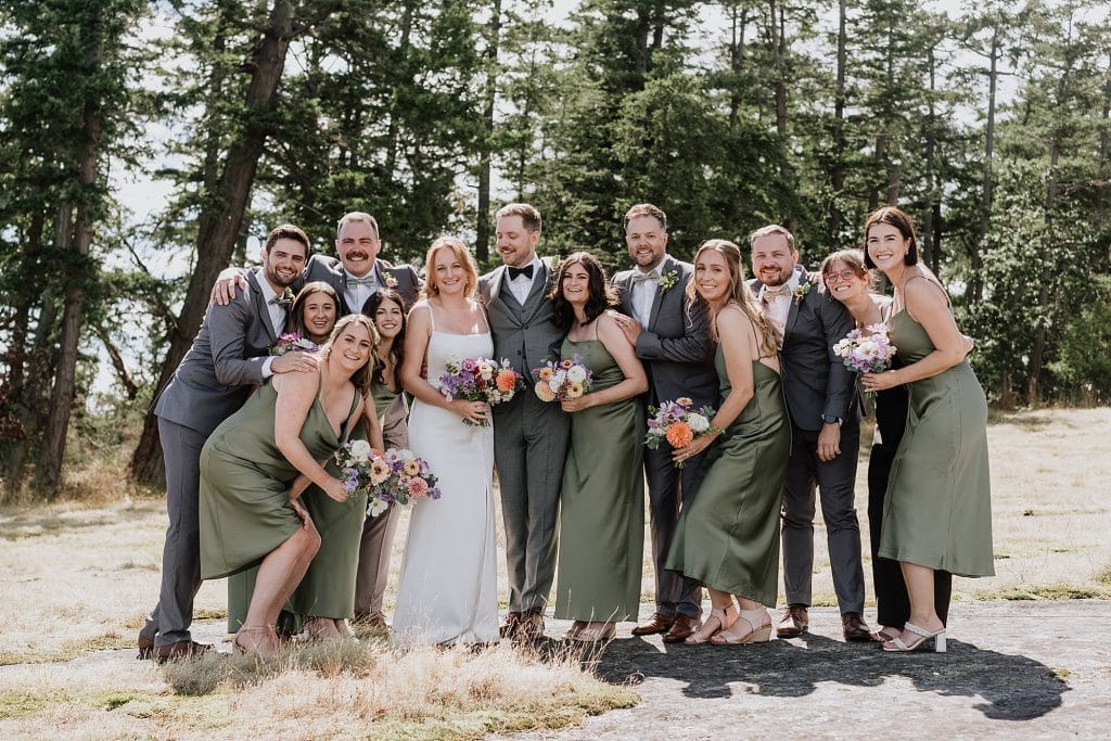 A wedding party poses outdoors, with the bride in white, groom in gray, bridesmaids in green dresses, and groomsmen in gray suits, all holding flowers and smiling in front of trees.