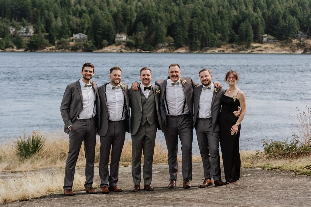 Six people stand together outdoors by a lake, dressed in formal attire, with a forested hillside visible across the water in the background.