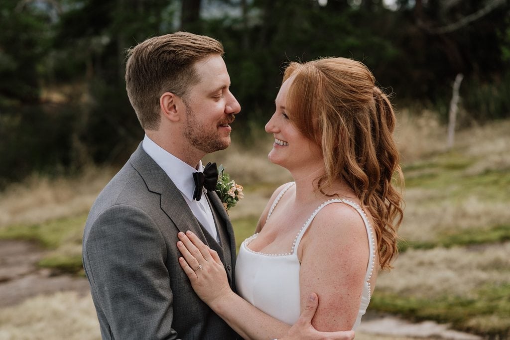 A couple dressed in wedding attire stands outdoors, facing each other and smiling, with greenery and dry grass in the background.