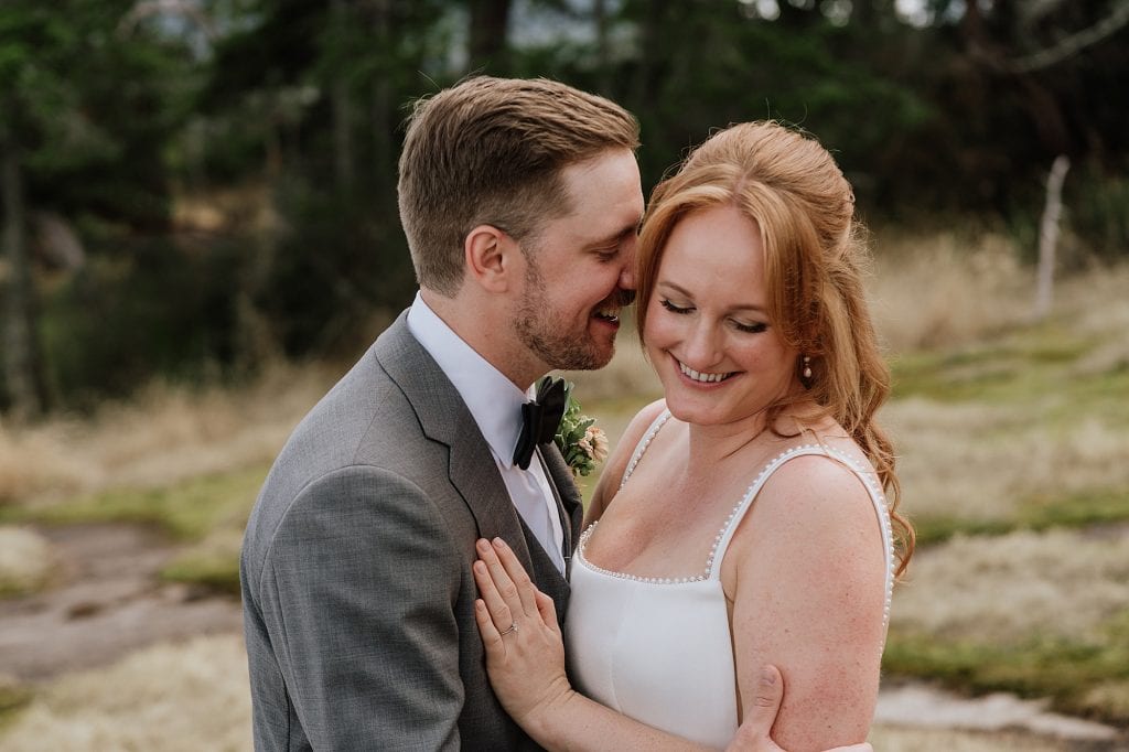 A man in a grey suit embraces a woman in a white dress outdoors. Both are smiling, and the background features grass and trees.