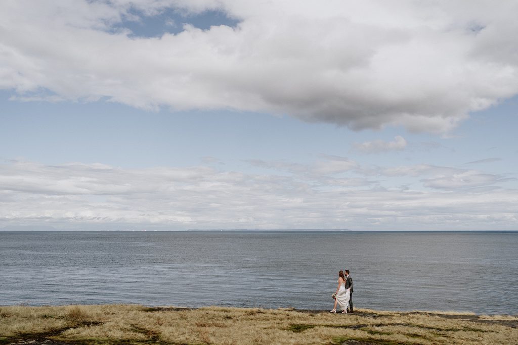 A couple stands together on a grassy coastline under a cloudy sky, with the ocean stretching out behind them.