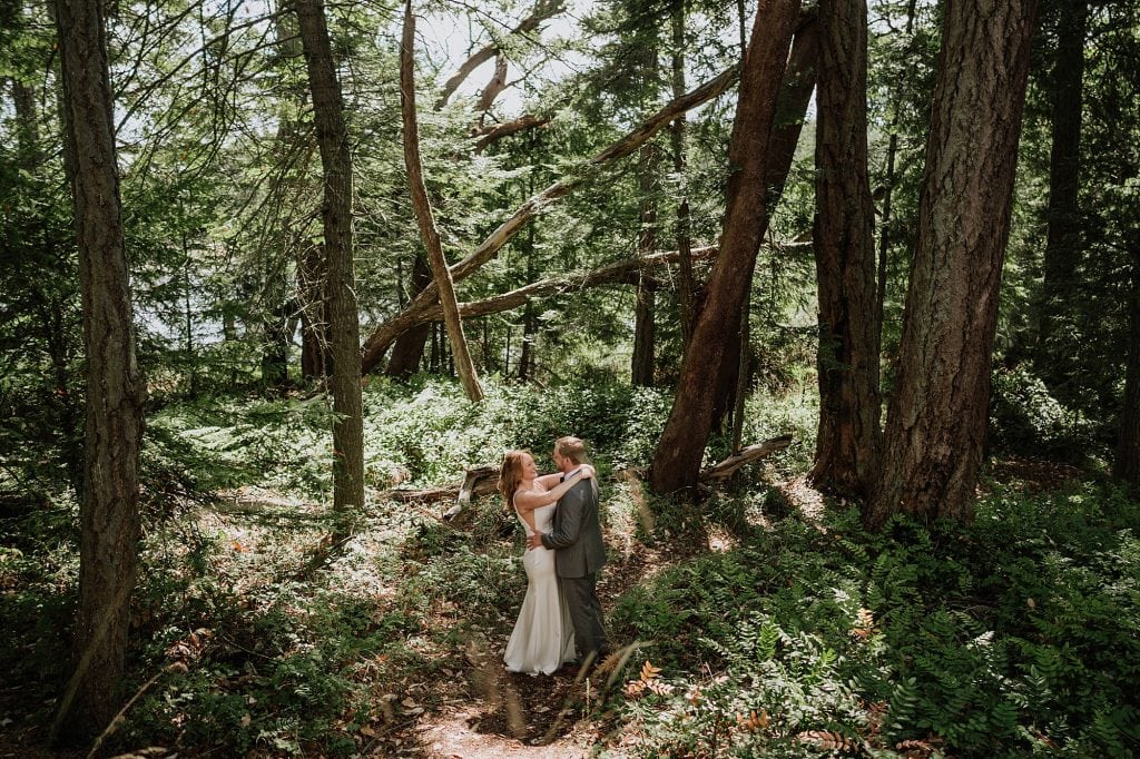 A couple in wedding attire embraces on a forest path surrounded by tall trees and greenery.