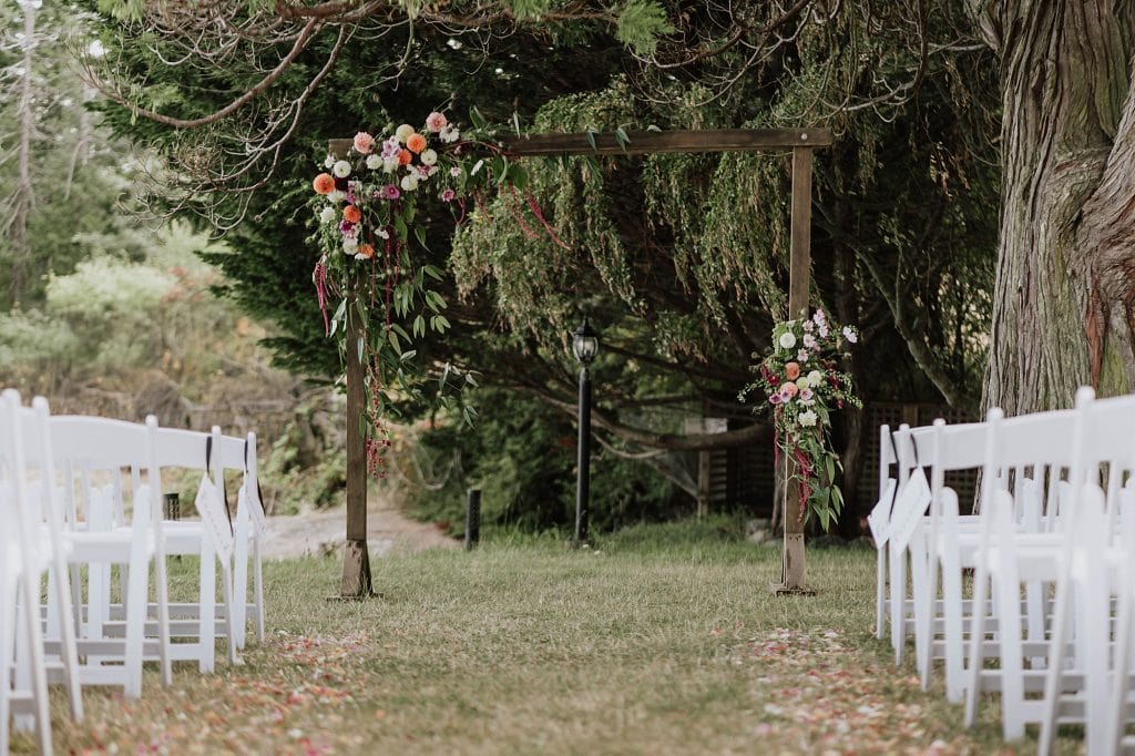 A wooden wedding arch decorated with flowers stands at the end of an aisle lined with white chairs on grass, set outdoors among trees.