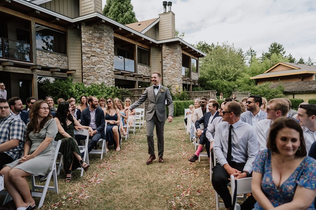 A man in a gray suit walks down the aisle outdoors between seated guests at a wedding ceremony, with a stone building in the background.