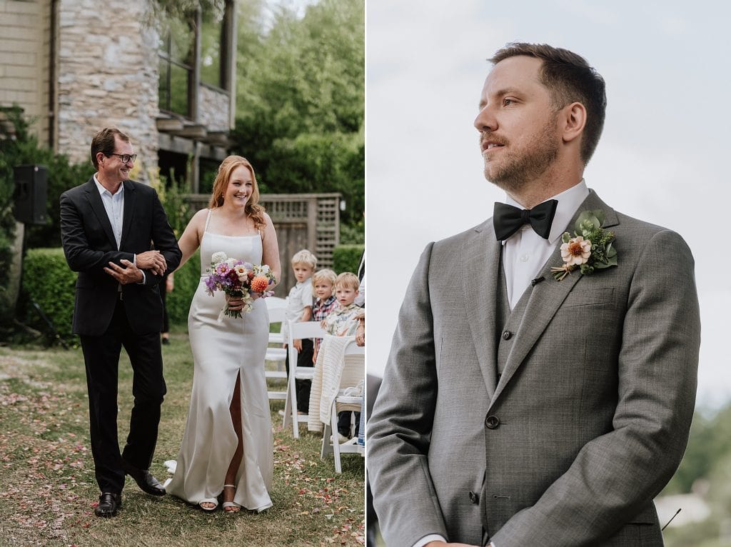 A bride in a white dress walks down the aisle with a man, while a groom in a gray suit waits with a boutonniere, during an outdoor wedding ceremony.