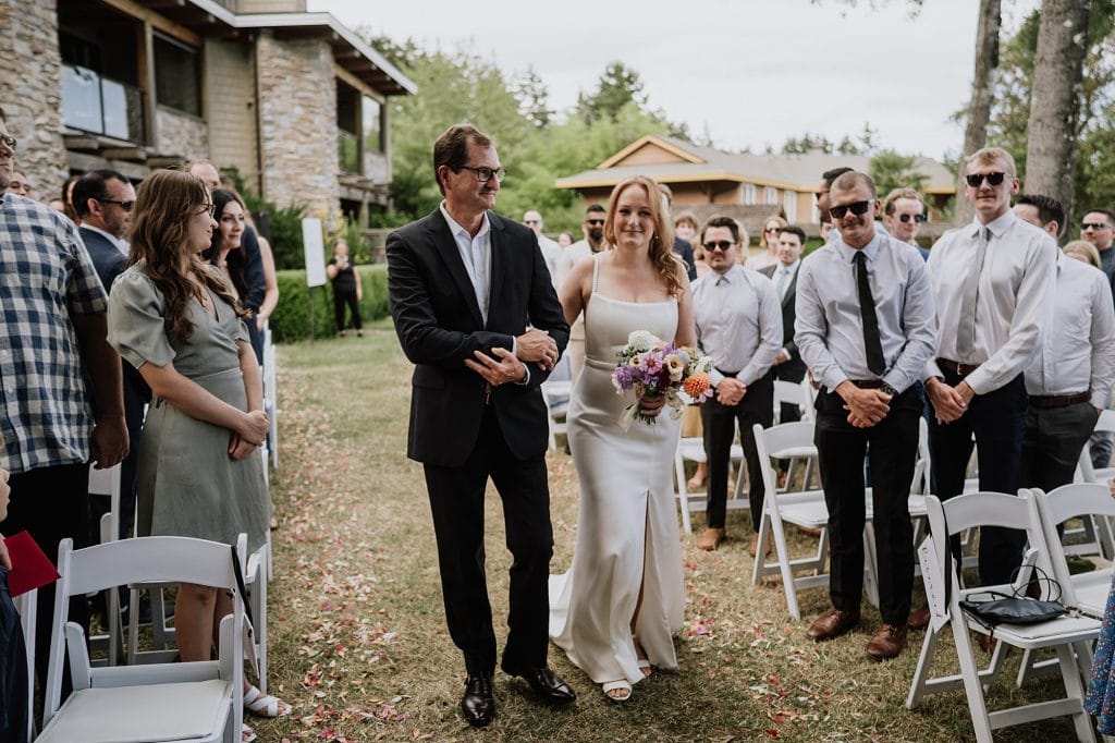 A bride in a white dress walks down the aisle with a man in a suit, holding a bouquet, as guests stand and watch at an outdoor wedding ceremony.