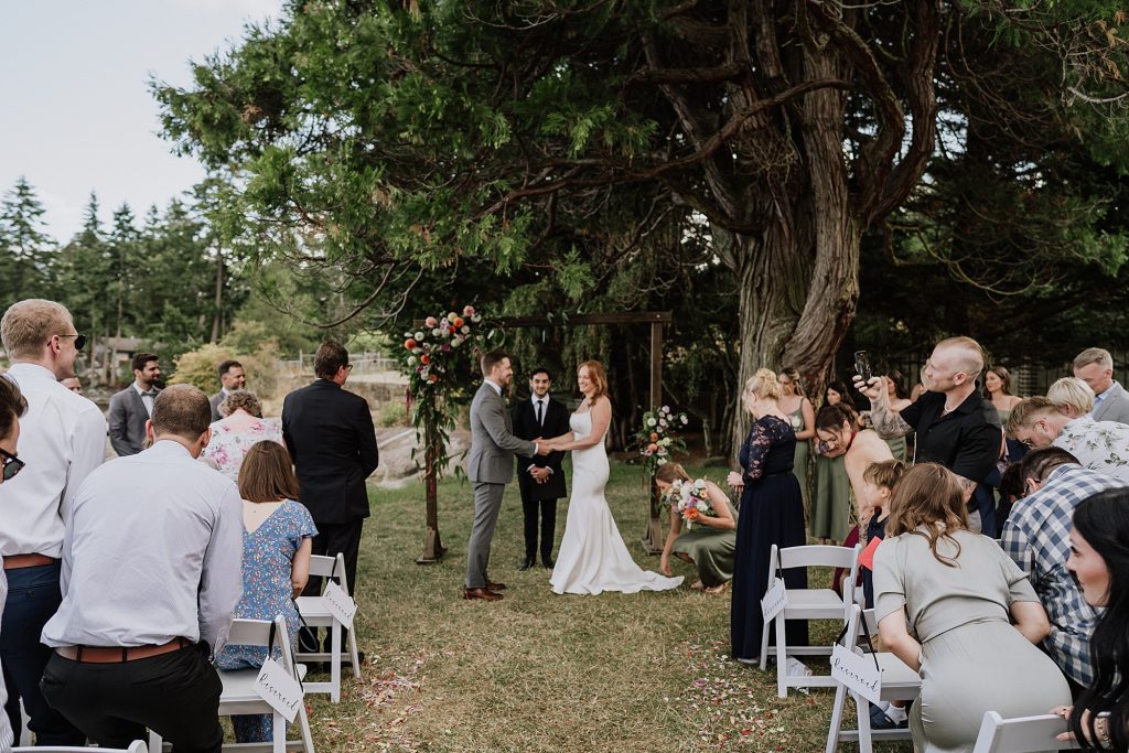 A bride and groom stand at an outdoor altar under a large tree during their wedding ceremony, surrounded by guests seated in white chairs.
