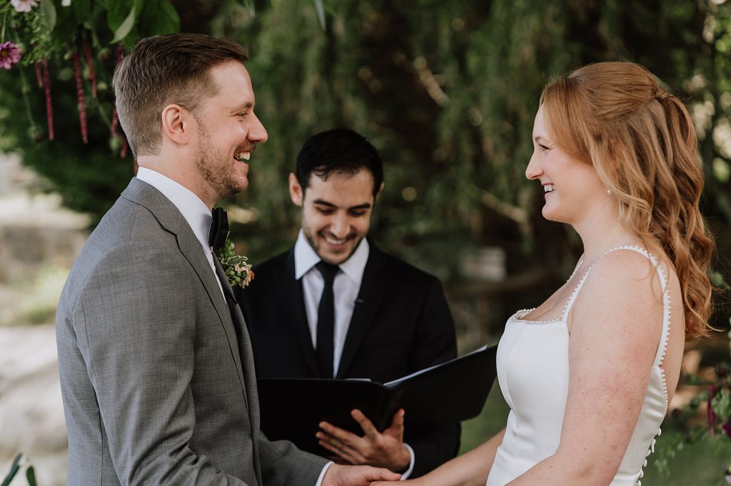 A man and woman stand facing each other, holding hands and smiling, while an officiant stands behind them with a book, outdoors.