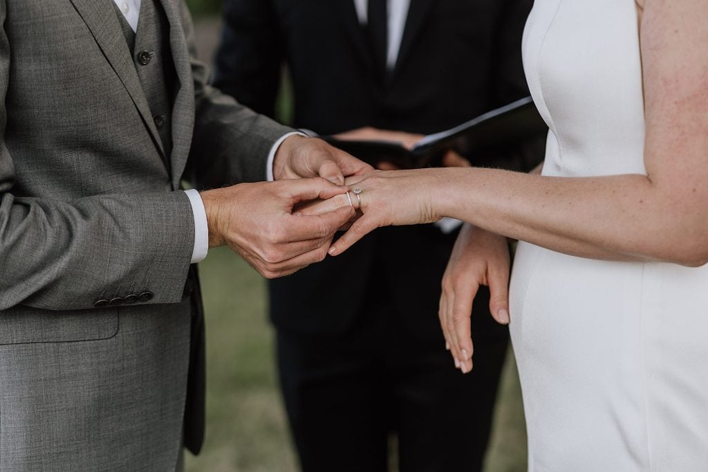 A person in a grey suit places a ring on the finger of a person in a white dress during a wedding ceremony, with an officiant standing in the background.