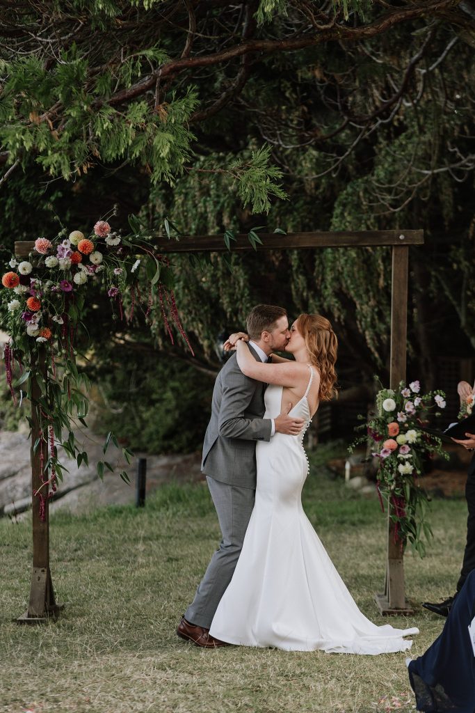 A bride and groom kiss under a wooden arch decorated with flowers during an outdoor wedding ceremony.