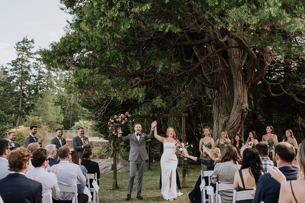 A bride and groom stand holding hands under a wooden arch during an outdoor wedding ceremony, surrounded by guests and a bridal party, with large trees in the background.