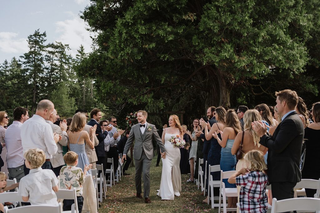 A bride and groom walk down an outdoor aisle lined with seated and standing guests applauding under large trees.