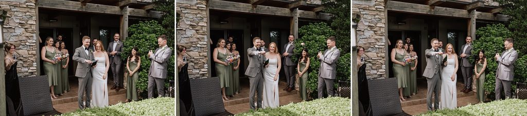 A bride and groom walk out of a stone building, followed by their wedding party. All are smiling and holding drinks, with greenery in the background.