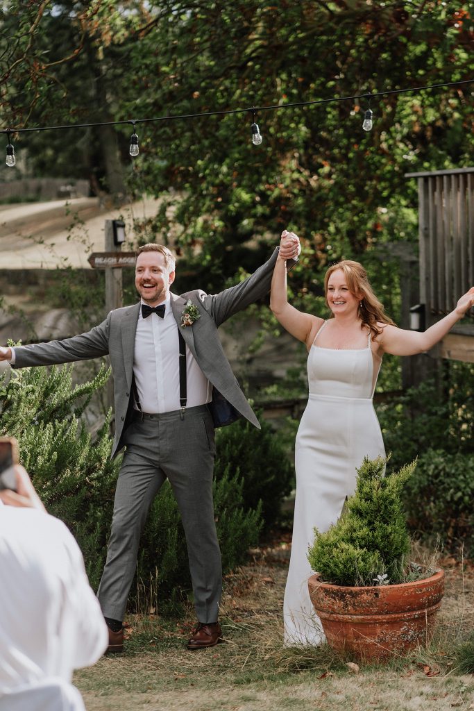A man in a gray suit and a woman in a white dress stand outdoors, holding hands and smiling, appearing to celebrate at an event.