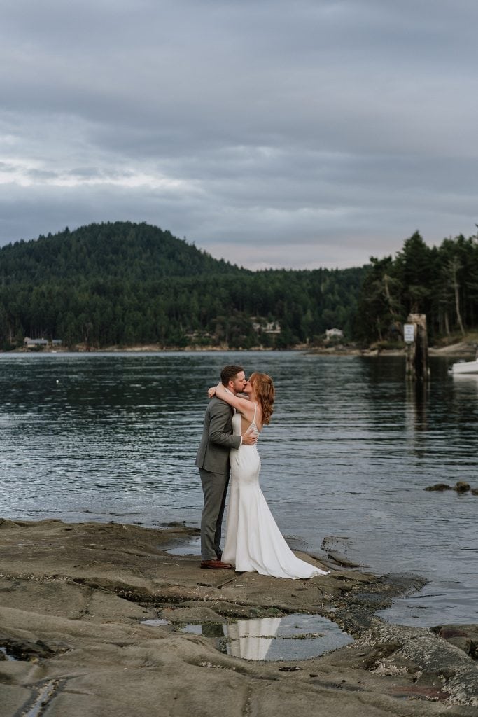 A couple dressed in wedding attire embrace on a rocky shore with a forested hillside and calm water in the background.
