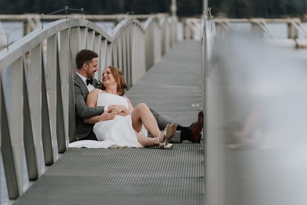 A bride and groom sit together on a metal bridge, smiling at each other, with the bride leaning back against the groom.