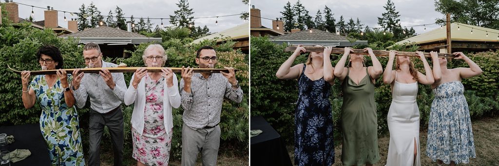 Two groups of people stand outdoors, each holding and drinking from a long wooden shot ski together. Shrubs and string lights are visible in the background.