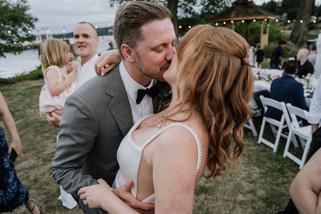 A bride and groom kiss outdoors at their wedding reception while guests, including a man holding a child, are visible in the background.