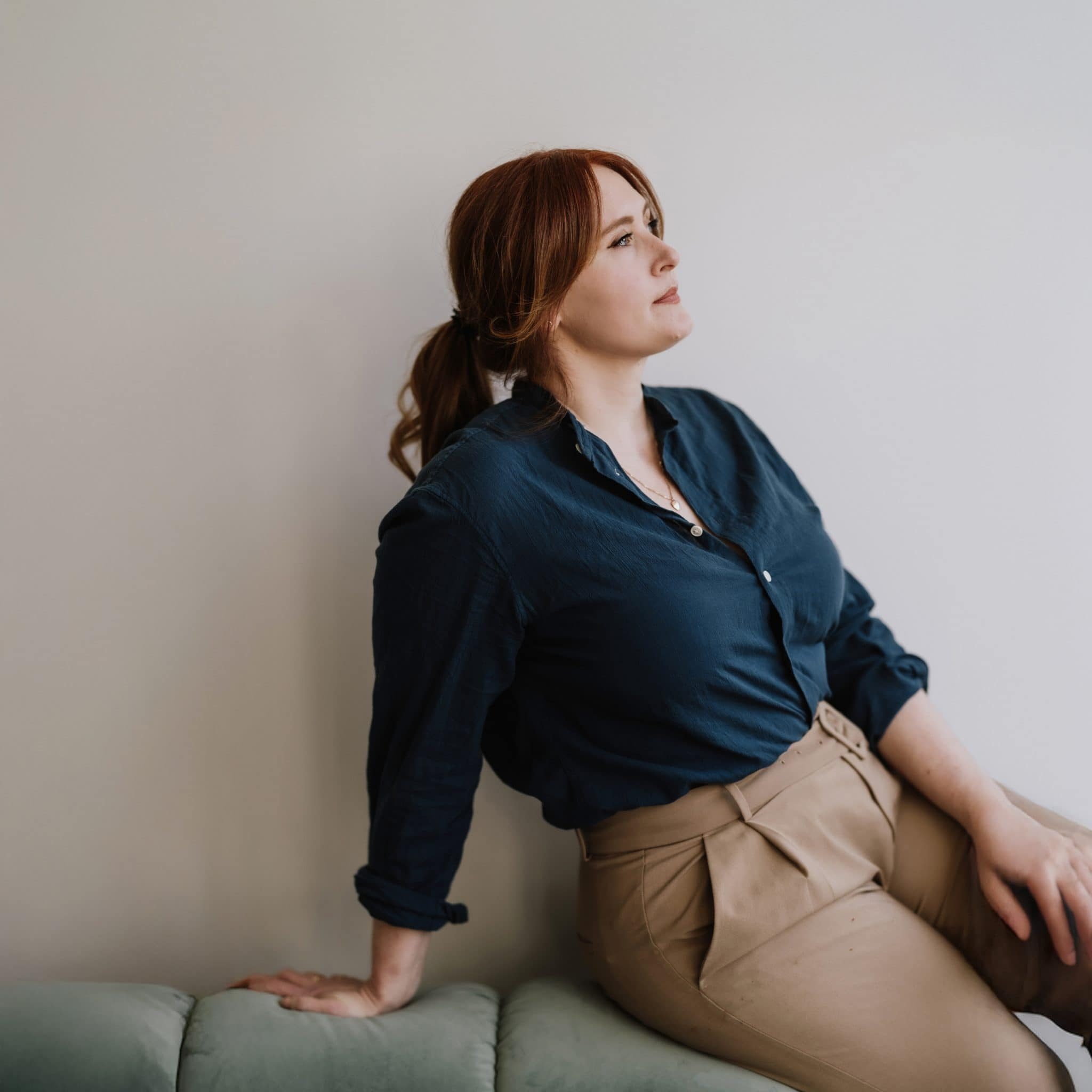 A woman with red hair in a ponytail, wearing a navy shirt and beige pants, sits on a green cushioned bench, looking to the side against a plain light-colored wall.
