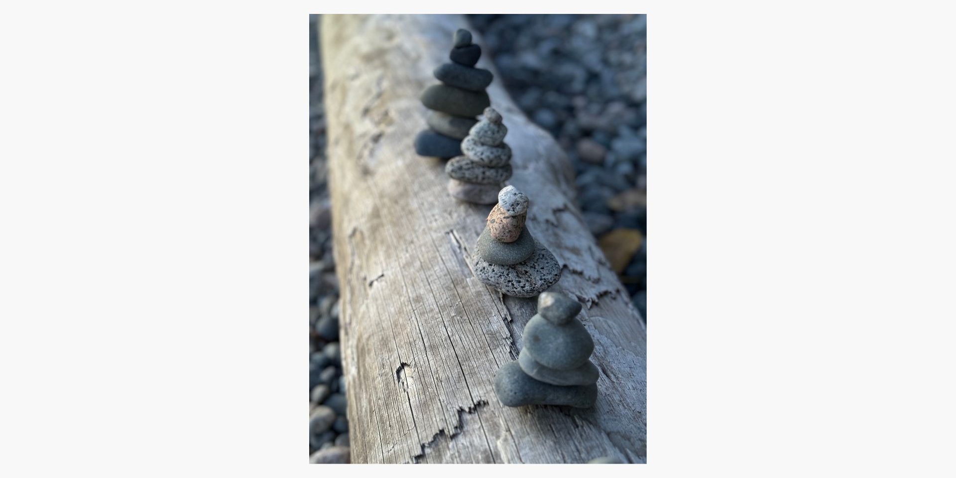 Several small stacks of balanced stones are arranged in a row on a weathered wooden log, with pebbles visible in the background.